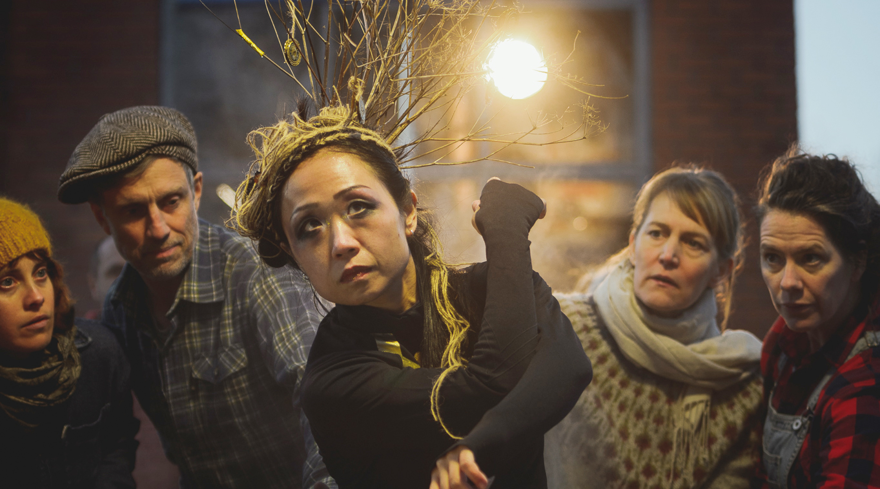 En grupp människor står på rad och tittar in i kameran. En stark lampa lyser i bakgrunden. En bild som syftar till att väcka uppmärksamhet och marknadsföra en föreställning.