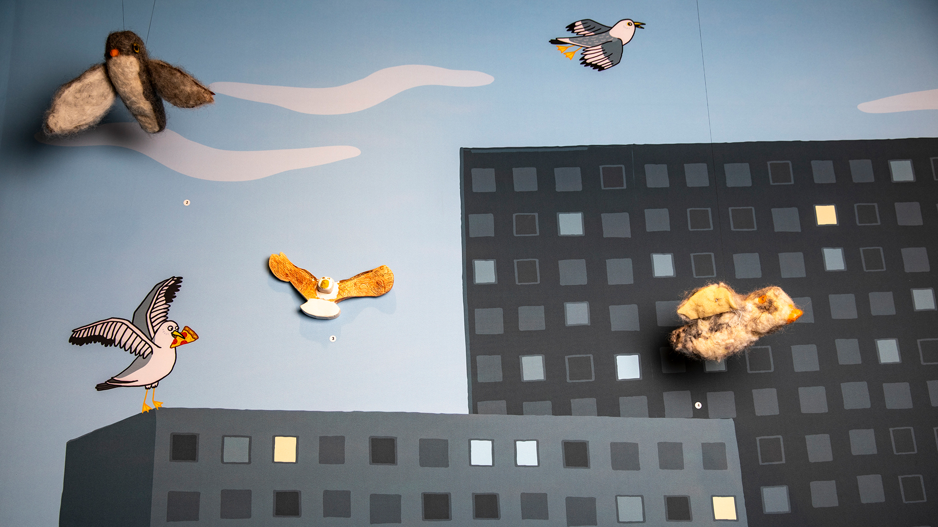 Två ulltovade fåglar skapade av barn hänger i trådar från taket mot en bakgrund med en illustratuon av höghus och måsar gjord av Sarah Sheppard. Även en mås av okänt material skapad av ett barn är uppsatt på väggen med illustrationen.