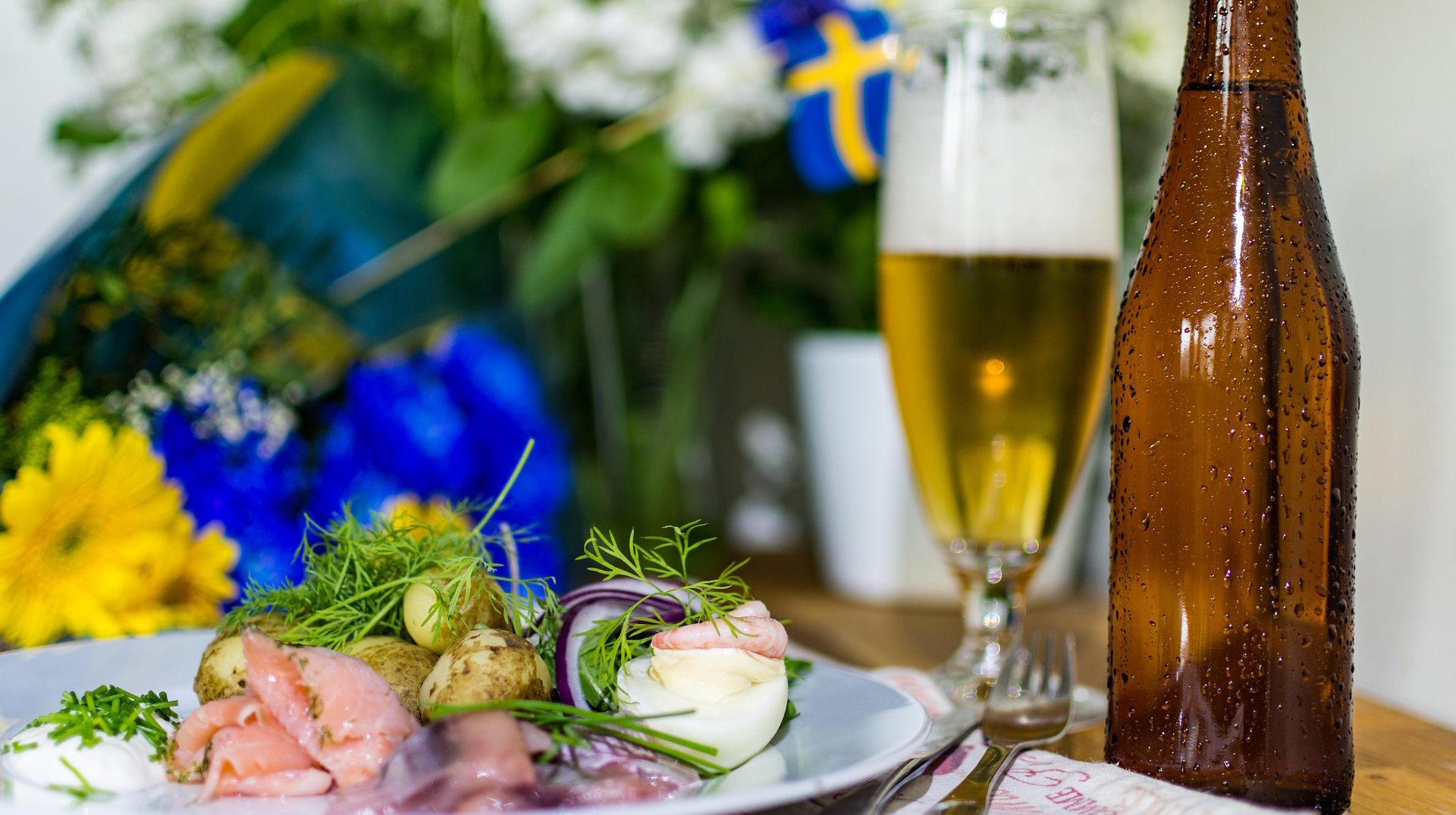 En uppdukad midsommarmåltid. En bild som illustrerar GNM:s arbete med biologisk mångfald. 
ALT TEXT: gula och blåa blommor, en liten svensk flagga. Till höger på bordet en brun ölflaska. Till vänster om den ett glas med skummande öl. Framför en silltallrik.