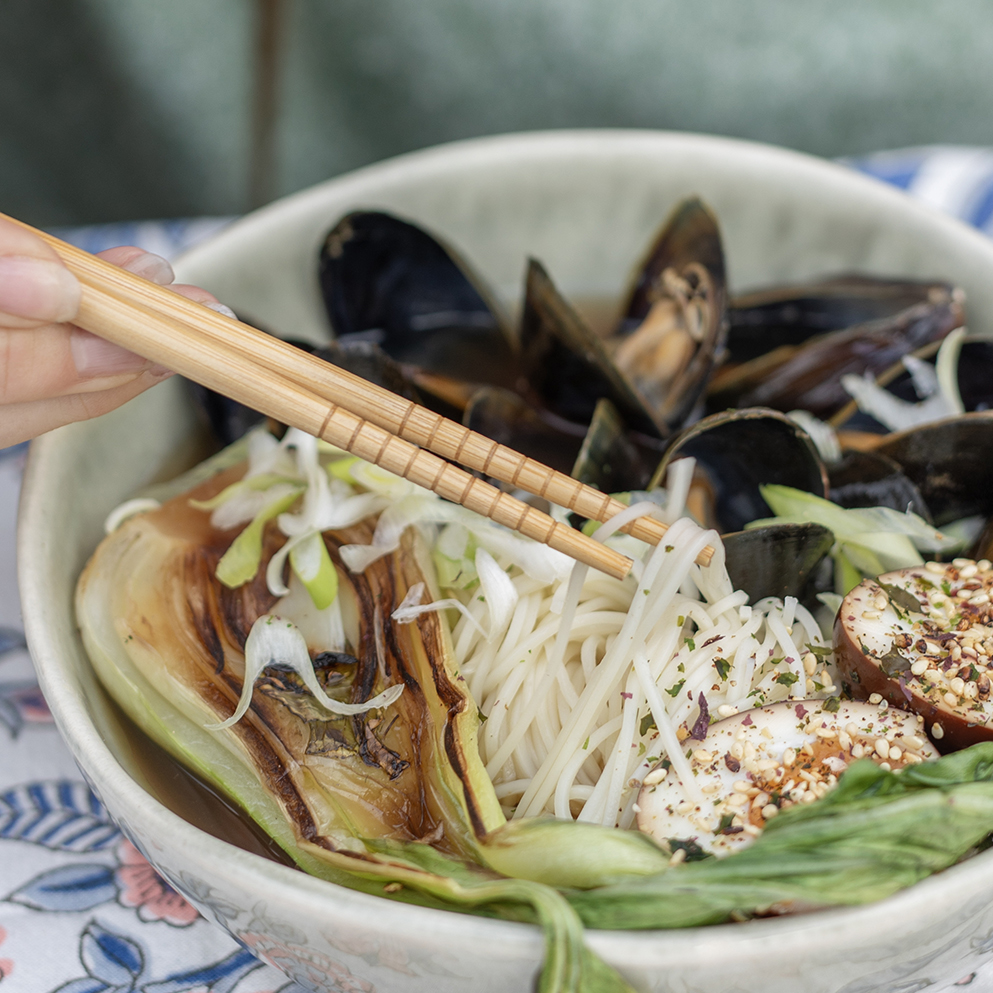 En portion ramen. En vit skål. Till väster ser vi en hand som håller i ett par ätpinnar som lyfter lite nudlar. I skålen ser vi även musslor, sesamfrön som lagts ovanför ägghalvor samt sallad. 