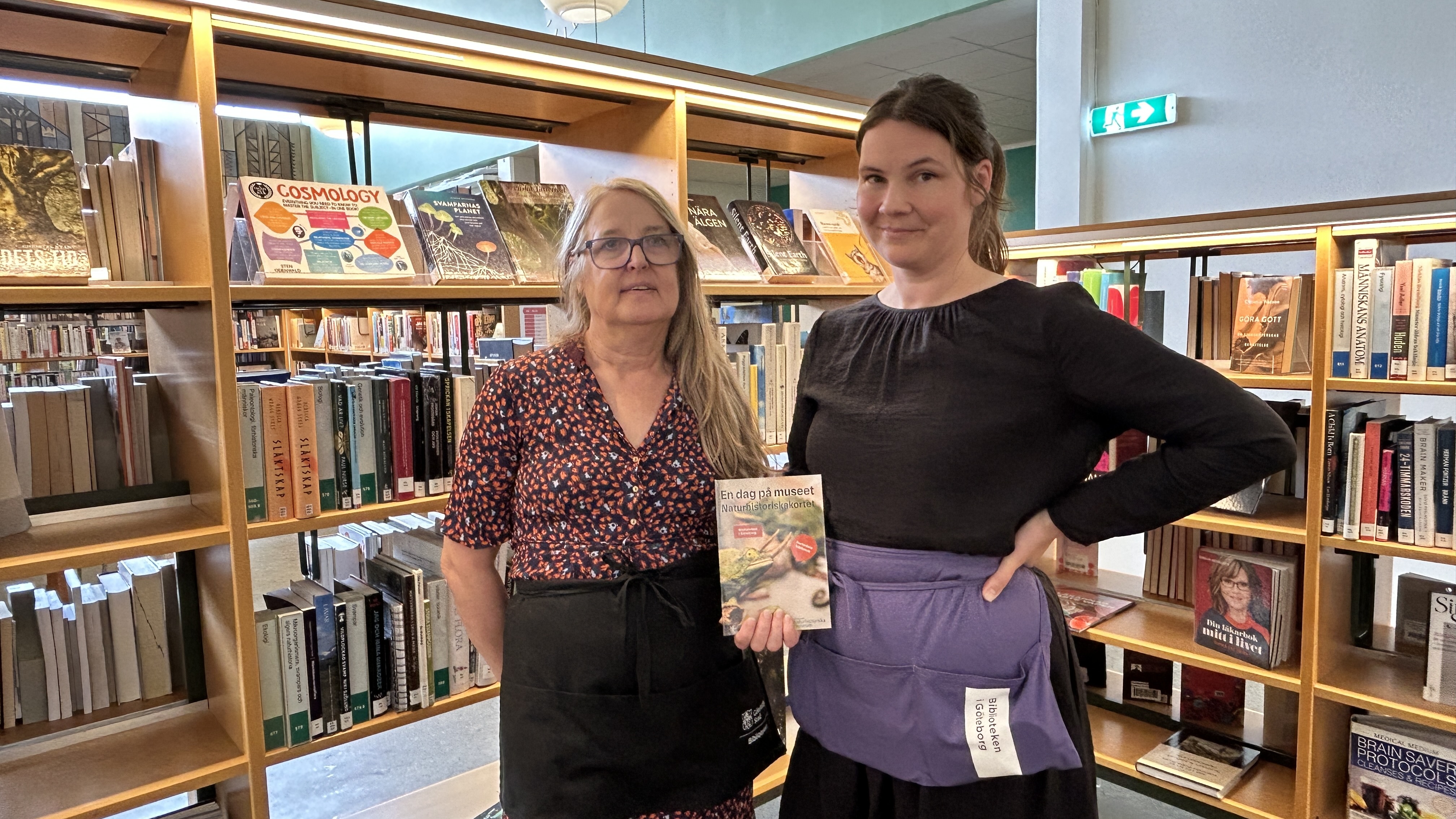 Två kvinnor står vid en bokhylla med böcker. Det är ett bibliotek. Kvinnan till vänster har långt, blont hår och glasögon. Hon har en mänstrad blus på sig i färgerna orange, svart och vitt. Hon har en svart kjol. Kvinnan till höger har mörkt hår och svart klänning. PÅ magen har hon ett lila, kort förkläde. Hon håller i en broschyr där Naturhistoriskakortet ligger.