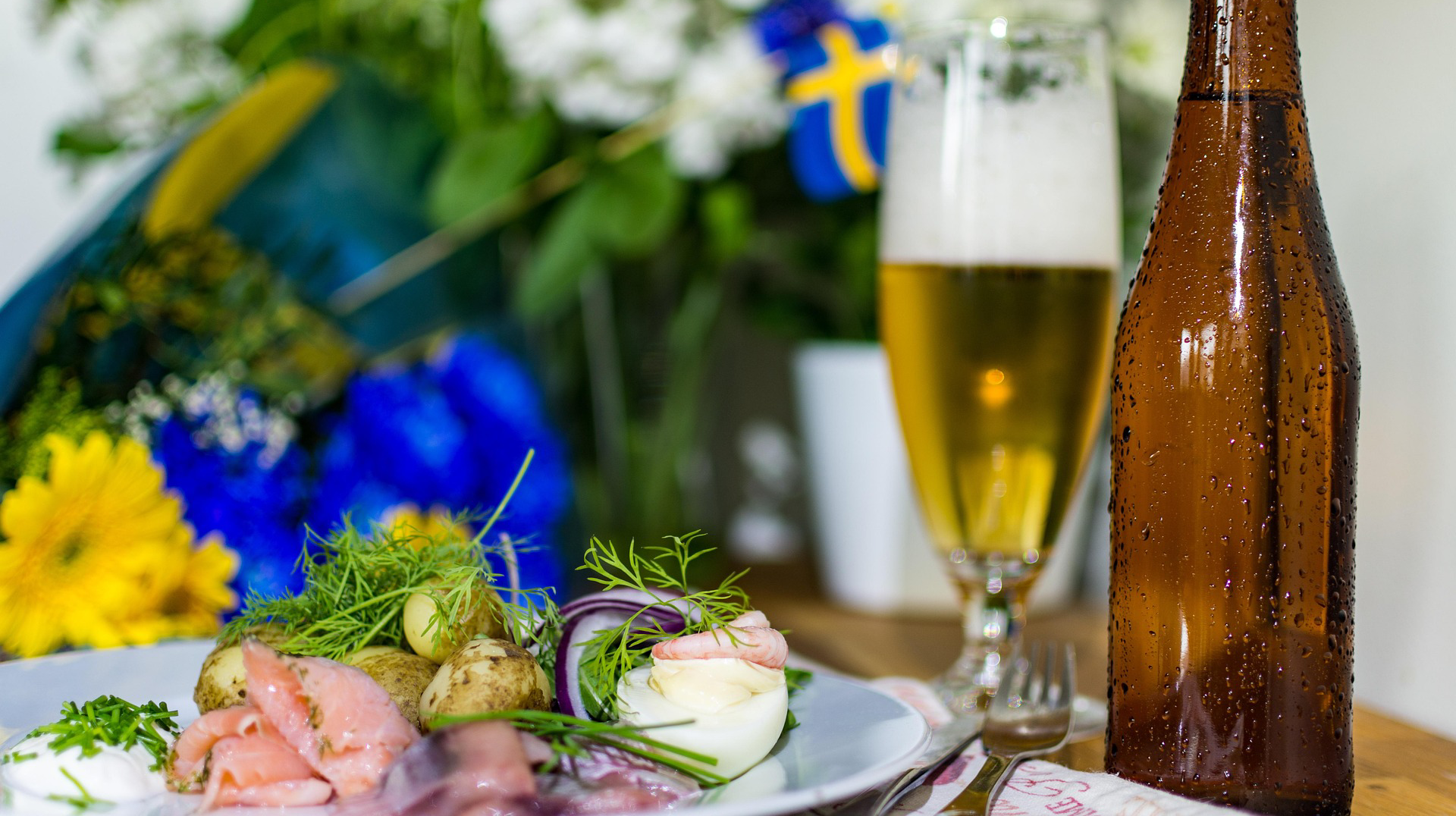 ula och blåa blommor, en liten svensk flagga. Till höger på bordet en brun ölflaska. Till vänster om den ett glas med skummande öl. Framför en silltallrik.
