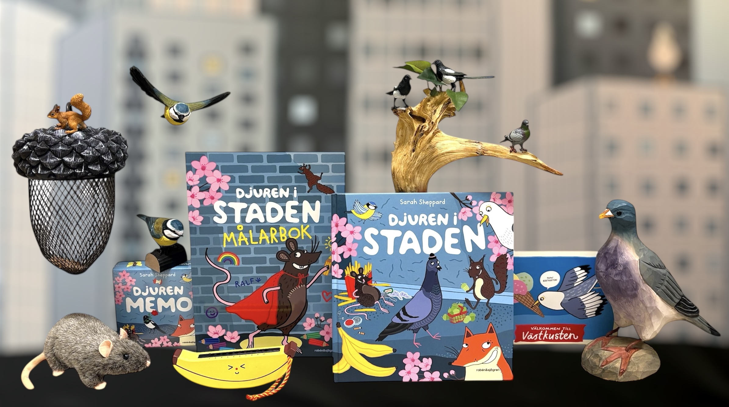 En samling varor från butiken på temat Djuren i staden (utställning) fotograferade mot en suddig bakgrund med en illustration av höghus. Bland annat böcker, snidade djur av trä och spel.
